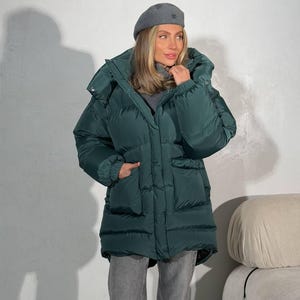 May include: A dark green puffer jacket with a large hood and pockets. The jacket is worn with a gray beret and gray pants. The jacket appears to be made of a quilted material.