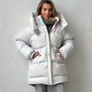 Puede incluir: Una chaqueta acolchada blanca con una gran capucha y bolsillos. La chaqueta se lleva sobre un cuello alto gris y se combina con vaqueros grises. La modelo está de pie frente a una pared blanca texturizada. La chaqueta parece cálida y cómoda.