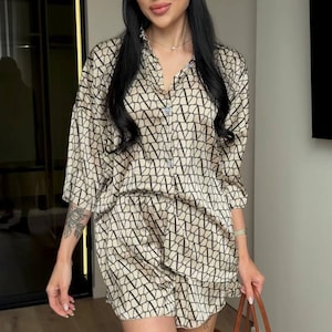Puede incluir: Un conjunto de dos piezas compuesto por una camisa abotonada y pantalones cortos de tela beige con un patr&oacute;n geom&eacute;trico repetitivo de letras V en negro. El conjunto incluye un pa&ntilde;uelo a juego, gafas de sol negras y un bolso de paja grande.