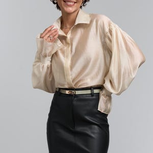 May include: A cream-coloured blouse with long, voluminous sleeves and a collared neckline. The blouse is paired with a black leather skirt and a thin belt with a gold buckle. The outfit is completed with glasses and earrings.