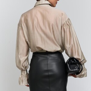 May include: A beige silk blouse with long sleeves and a black leather mini skirt. The blouse has a collared neckline and buttoned cuffs. A small black drawstring bag is held at the side.