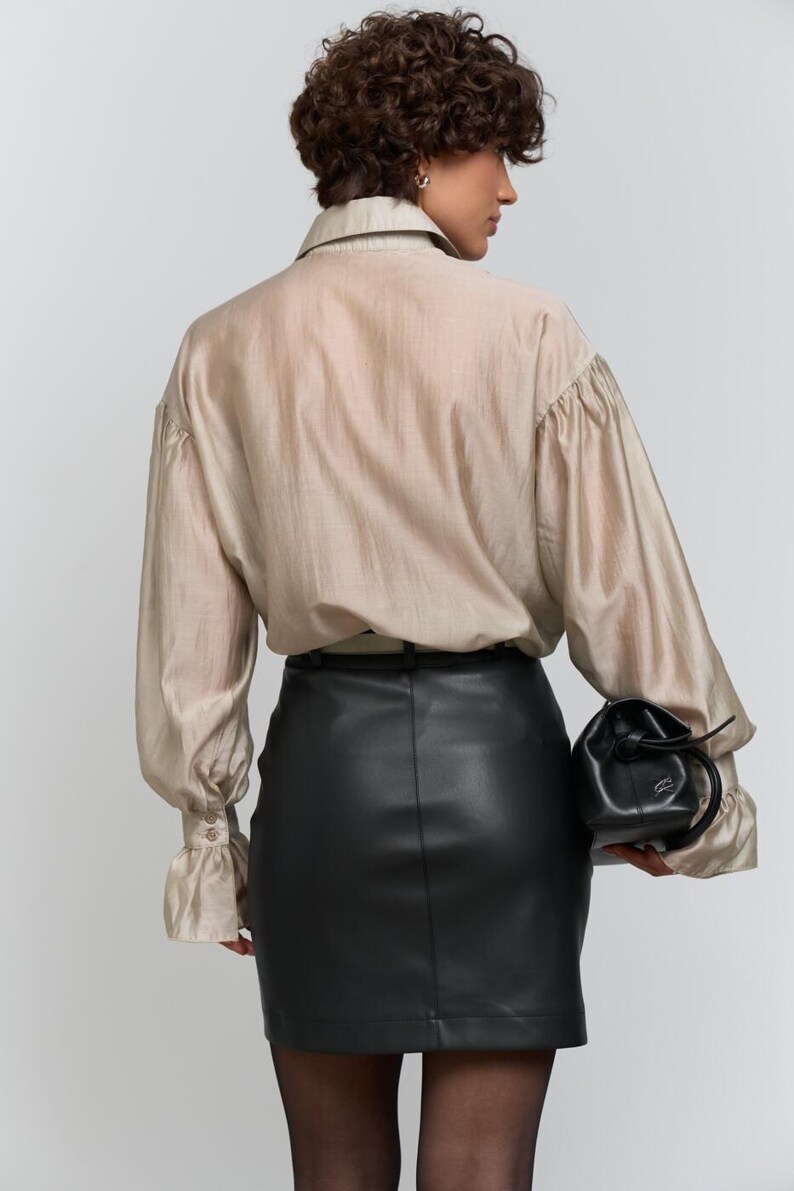 May include: A beige silk blouse with long sleeves and a black leather mini skirt. The blouse has a collared neckline and buttoned cuffs. A small black drawstring bag is held at the side.