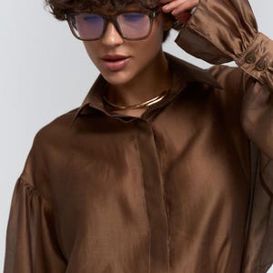 May include: A brown, sheer button-down shirt with long sleeves and a collar. The model is wearing square-framed glasses and a gold necklace. The shirt is paired with a light-coloured belt with a circular buckle.