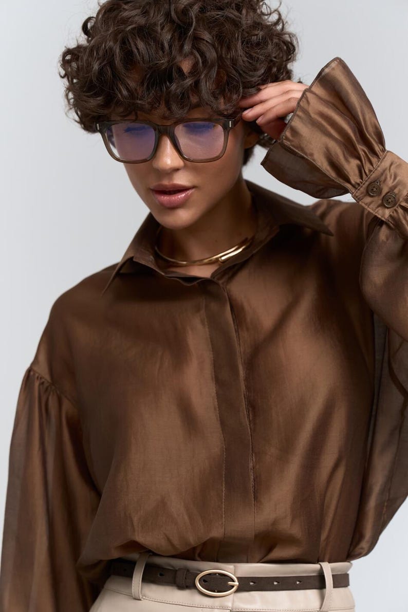 May include: A brown, sheer button-down shirt with long sleeves and a collar. The model is wearing square-framed glasses and a gold necklace. The shirt is paired with a light-coloured belt with a circular buckle.