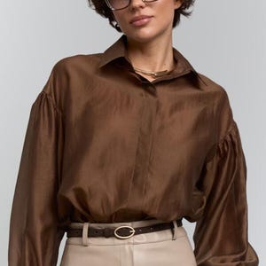 May include: A brown silk blouse with long, voluminous sleeves and a collared neckline. The blouse is paired with beige trousers and a brown belt with a gold buckle. A gold necklace and bracelet complete the look. The model is wearing glasses.