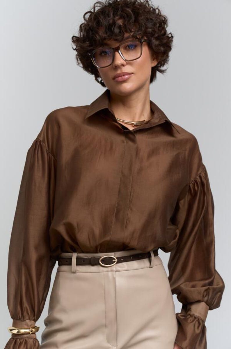 May include: A brown silk blouse with long, voluminous sleeves and a collared neckline. The blouse is paired with beige trousers and a brown belt with a gold buckle. A gold necklace and bracelet complete the look. The model is wearing glasses.