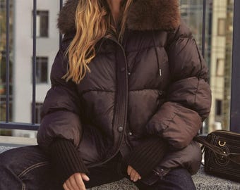 May include: A brown puffer jacket with a faux fur collar. The jacket has a button-up front and ribbed cuffs. The person is wearing dark sunglasses and dark wash jeans. A black purse is on the right.