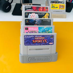 Può includere: Un espositore a gradini in plastica grigia contenente cinque cartucce di gioco Super Famicom. Il supporto presenta il logo Nintendo in rilievo sulla parte anteriore. Le cartucce includono titoli come Samurai Spirits, Super Mario e altre etichette.