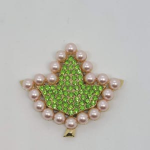 May include: A gold-toned brooch in the shape of a leaf. The leaf's center is filled with small, sparkling green rhinestones. The leaf is surrounded by a border of pink pearls. The brooch has a gold stem.