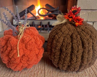 May include: Two round, plush, crocheted pumpkins. The left pumpkin is bright orange with a stem wrapped in twine and topped with sprigs of dried lavender. The right pumpkin is dark brown, decorated with a burlap bow and small red and orange flowers.