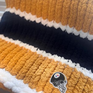 May include: A chunky knit blanket featuring horizontal stripes in mustard yellow, black, and white. The texture is thick and soft, created with large yarn loops. A patch depicting a black football helmet with a logo is attached to the surface.