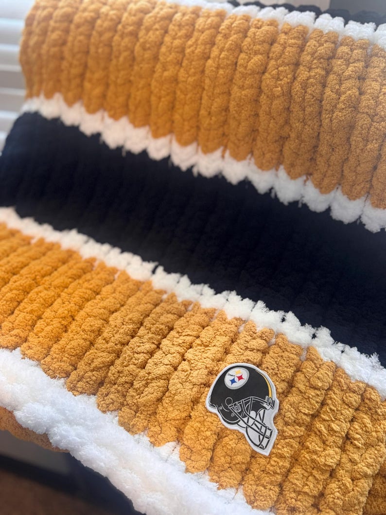 May include: A chunky knit blanket featuring horizontal stripes in mustard yellow, black, and white. The texture is thick and soft, created with large yarn loops. A patch depicting a black football helmet with a logo is attached to the surface.