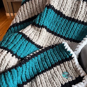 May include: A chunky knit throw blanket featuring horizontal stripes in teal, beige, and black. The texture is thick and plush, created with large yarn loops. A small embroidered football helmet patch in teal, black, and white is visible on the corner.