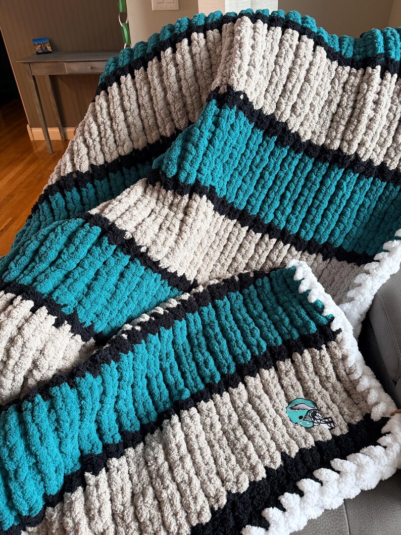 May include: A chunky knit throw blanket featuring horizontal stripes in teal, beige, and black. The texture is thick and plush, created with large yarn loops. A small embroidered football helmet patch in teal, black, and white is visible on the corner.