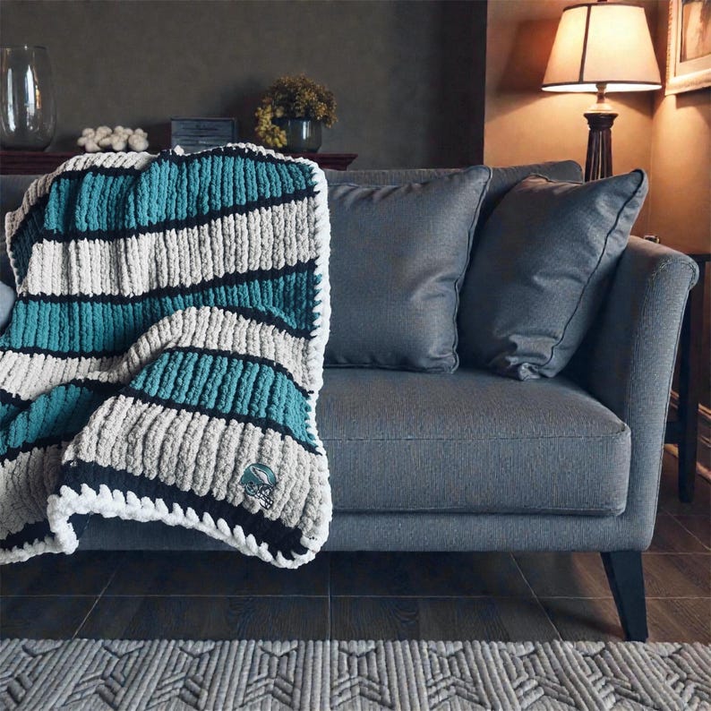 May include: A chunky knit throw blanket featuring horizontal stripes in teal, cream, and black. The blanket has a thick, textured yarn construction with a small embroidered logo patch near the bottom corner, draped over a gray upholstered sofa.