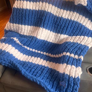 May include: A chunky knit throw blanket featuring horizontal stripes in royal blue and white. The blanket has a thick, soft texture and includes a small, embroidered blue lion logo patch positioned near the bottom right corner of the fabric.