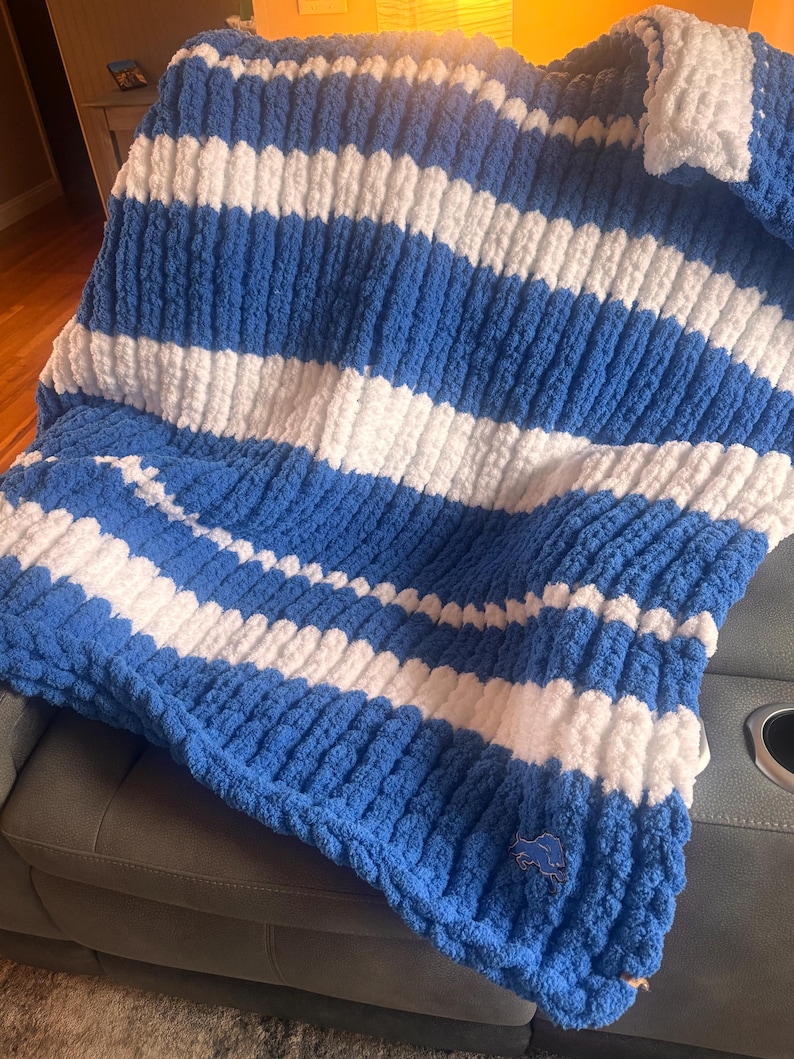 May include: A chunky knit throw blanket featuring horizontal stripes in royal blue and white. The blanket has a thick, soft texture and includes a small, embroidered blue lion logo patch positioned near the bottom right corner of the fabric.