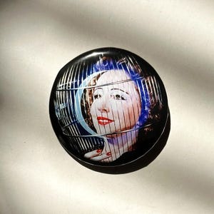 May include: A black and white button featuring a woman's face within a cage-like structure. The woman is wearing red lipstick and blue eyeshadow. The background is black.