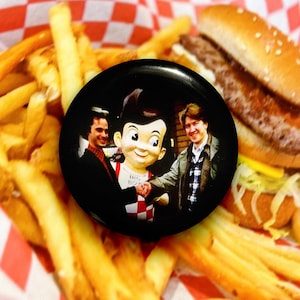 Op de afbeelding: Een zwarte knoop met het logo van het Big Boy restaurant, met twee mannen die de hand schudden van de Big Boy mascotte. De knoop staat tegen een achtergrond van friet, een hamburger en rood-wit geruit papier.