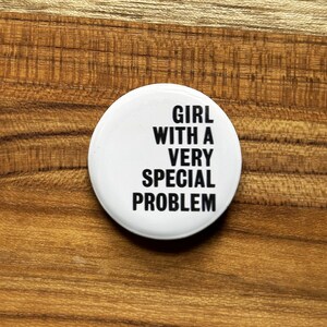 May include: A white button with black text that reads "GIRL WITH A VERY SPECIAL PROBLEM." The button is circular and sits on a wooden surface.
