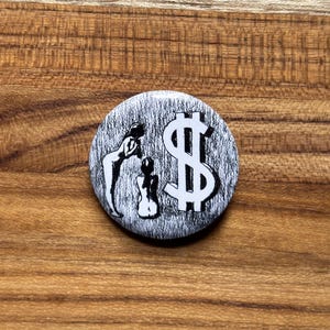 May include: A round button with a black and white design. The button features a dollar sign and two silhouetted figures. The background has a textured, gray appearance.