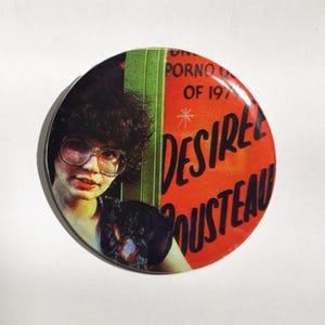 May include: Round button with a vintage design. The button features a woman with glasses and text that reads "PORNO OF 197-" and "DESIREE ROUSTEAU" in red and white. The background is orange and green.