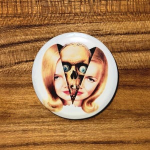 May include: A white button with a vintage-style image. The design features a skull bisecting the faces of two women with blonde hair and red lipstick. The skull has blue eyes. The button is round and has a glossy finish.