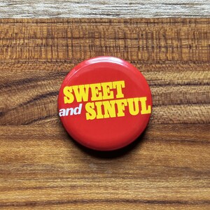 May include: A round, red button with the words "SWEET and SINFUL" in bold, yellow letters. The button is set against a wooden background.