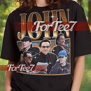 Op de afbeelding: Zwart unisex T-shirt met een grafische print van een band genaamd "John For Tee7". De afbeelding toont een groep mannen in een bandsetting met een vintage esthetiek.