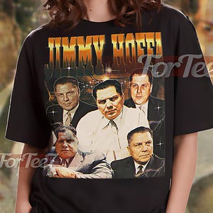 Puede incluir: Camiseta unisex negra con un gráfico de Jimmy Hoffa y otros hombres con trajes. La camiseta tiene el texto "JIMMY HOFFA" en letras doradas. La camiseta la lleva una persona.