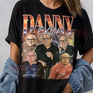May include: Black unisex t-shirt featuring a graphic of Danny DeVito in various poses, with the name "DANNY DeVito" in an orange and gold design. The shirt has the text "UNISEX SHIRT" at the top.