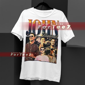May include: White t-shirt with a graphic print of a group of men with the word "JOHN" in large letters above them. The text "Original design by ForTee7" is printed in red and white on the top and bottom of the graphic.