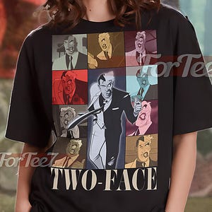 May include: Black unisex t-shirt featuring a graphic of the Two-Face character from DC Comics. The design includes multiple panels showcasing the character's split appearance, with the text "TWO-FACE" below. The shirt is labeled "UNISEX SHIRT".