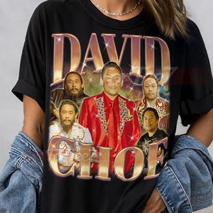 May include: Black unisex t-shirt featuring a retro-style graphic with the name "DAVID CHOE" in gold lettering. The design includes multiple images of a man in a red jacket. The shirt is a classic crew neck.