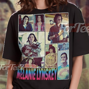 May include: Black unisex t-shirt featuring a collage of images and the name "MELANIE LYNSKEY" in pink and blue. The shirt has the text "UNISEX SHIRT" at the top. The model is wearing an XL shirt.