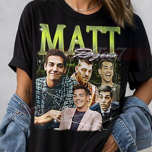 Peut inclure: T-shirt unisexe noir avec un graphisme de Matt Rogers et le texte "MATT Rogena". Le design comprend plusieurs images de Matt Rogers avec des éclairs. Le t-shirt a un col rond classique.
