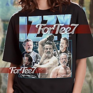 May include: Black unisex t-shirt with a graphic print of a man with a beard and a shirtless man holding a weapon. The text "IZZY For Tee7" is printed on the shirt.
