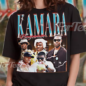 May include: Black unisex t-shirt featuring a graphic of a group of people with the word "KAMARA" in a blue and white font. The word "Benjamin" is below the main text. The shirt has a vintage style.