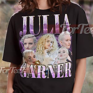 以下が含まれることがあります： 黒色のユニセックスTシャツ。紫と白のグラフィックでジュリア・ガーナーと「JULIA GARNER」の文字が描かれています。Tシャツの上部には「UNISEX SHIRT」の文字。モデルはXLサイズのシャツを着用しています。