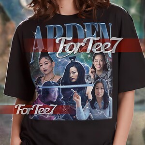 May include: Black unisex t-shirt featuring a graphic print of a group of women with the text "ARDEN For Tee7" in blue and red. The shirt is a relaxed fit and has short sleeves.
