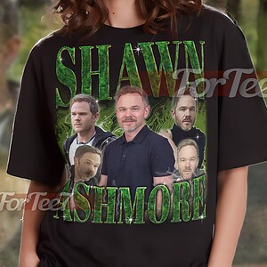 May include: Black unisex t-shirt with a green graphic of Shawn Ashmore and the name "SHAWN ASHMORE". The shirt has a crew neck design.