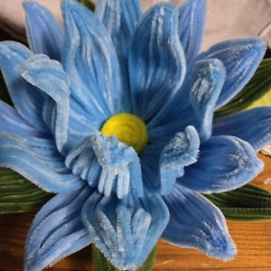 Op de afbeelding: Een blauwe viltbloem met een geel hart en groene bladeren. De bloem is gemaakt van chenille draad en is een knutselproject.