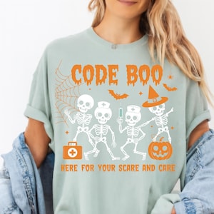 May include: A light green t-shirt with the words "CODE BOO" in orange, along with a spiderweb, bats, and skeleton figures. The phrase "HERE FOR YOUR SCARE AND CARE" is printed below the skeletons. The design is Halloween-themed.