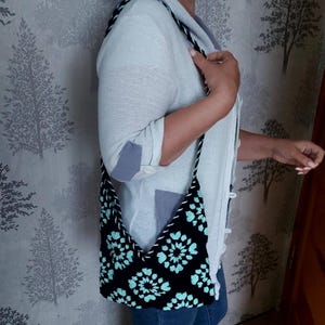 May include: Black shoulder bag with a turquoise floral pattern. The bag has a black and white rope strap. The bag is made of a soft material. The bag is worn over the shoulder.