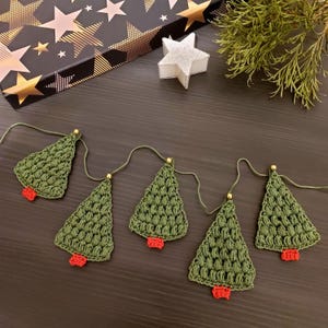 May include: A green crocheted Christmas tree garland with red ornaments. The garland is strung together with gold beads and white string.