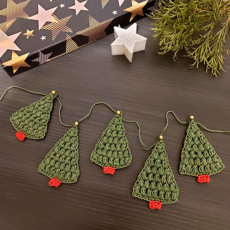 May include: A green crocheted Christmas tree garland with red ornaments. The garland is strung together with gold beads and white string.