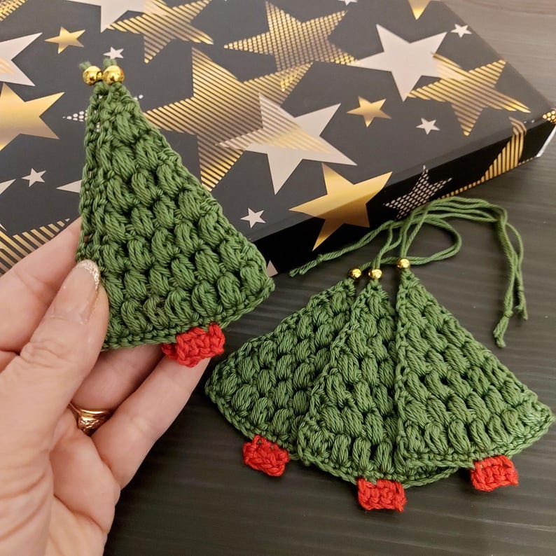 May include: Four green crocheted Christmas tree ornaments with red felt bases and gold beads on the top. The ornaments are hanging from a gold string.