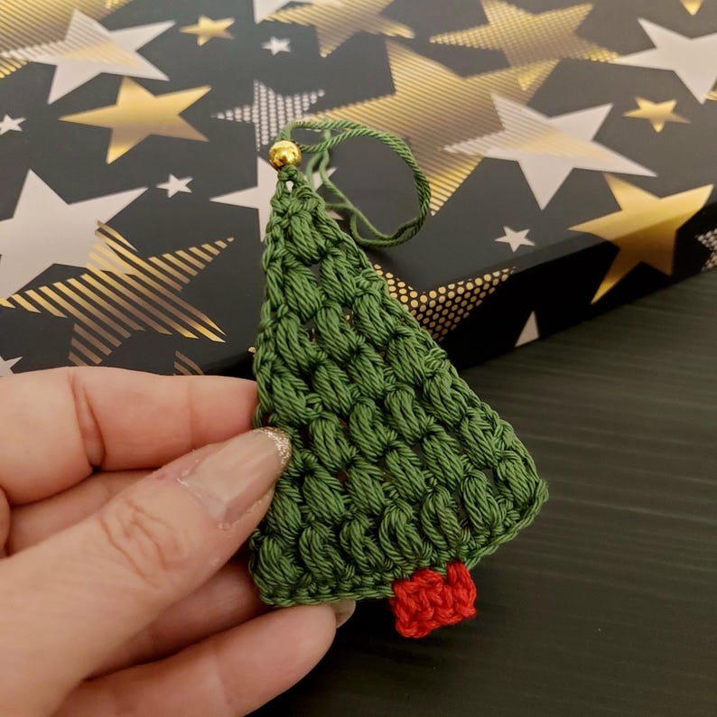 May include: A green crocheted Christmas tree ornament with a red felt star on top. The ornament is held in a hand against a background of gold stars.
