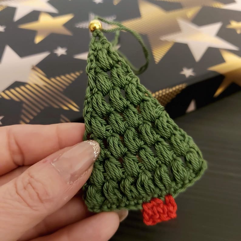 May include: A green crocheted Christmas tree ornament with a red felt base. The ornament has a gold bead at the top.