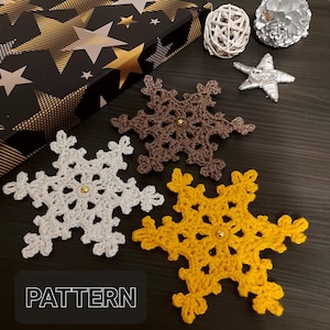May include: Three crocheted snowflakes in white, brown, and yellow. The snowflakes are on a dark wood surface with a gold and black gift box in the background. The text "PATTERN" is printed on the bottom of the image.